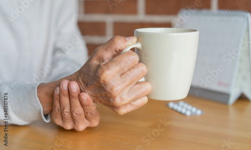 close up senior woman hold on hand to relief shaky symptom while drink water for Parkinson’s disease and chronic illness health care concept