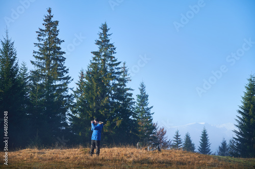 Male photographer hiker standing on grassy hill and taking picture with professional camera under blue sky