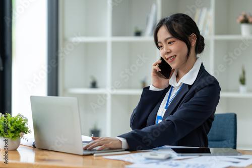 Asian businesswoman using mobile phone to communicate, chat, negotiate, search internet, news, email while working online on laptop computer at office.