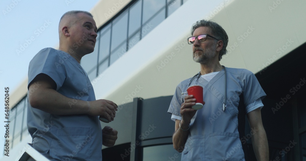 Obraz premium Two doctors in uniform stand near medical center entrance in the evening. Professional medics drink coffee during break and talk. Medical staff of hospital or modern clinic. Lifestyle and healthcare.