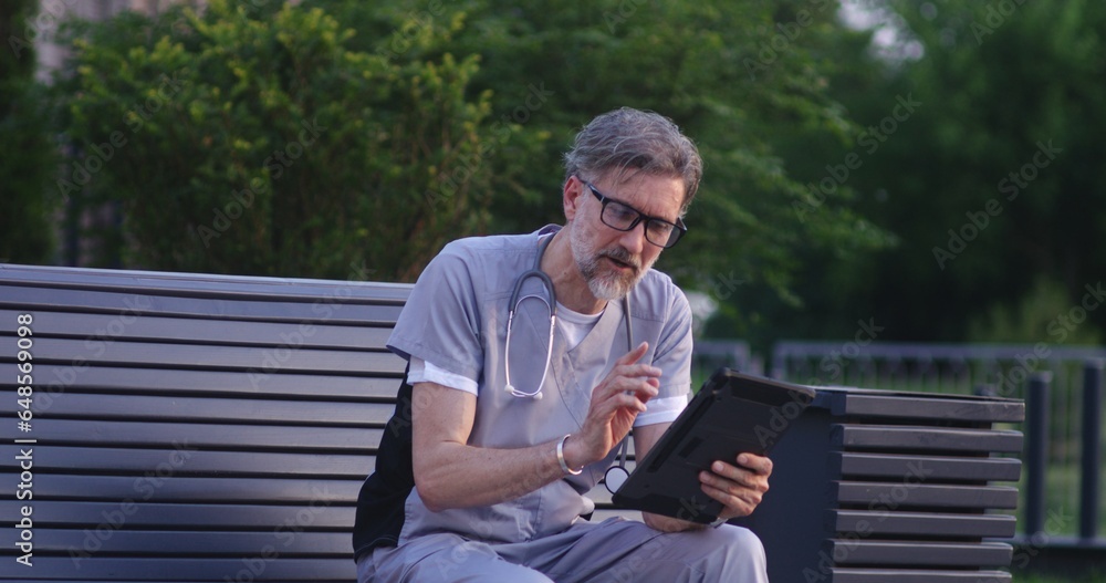 Fototapeta premium Doctor in uniform sits on bench outside modern hospital, talks to patient about treatment during online appointment using digital tablet. Medical staff of clinic or medical center. Healthcare system.