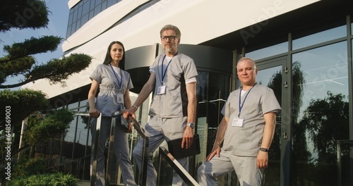 Diverse doctors in uniforms stand outdoors at the hospital entrance. Professional medics and nurse smile, look at camera. Medical staff of modern clinic or medical center. Healthcare system concept.