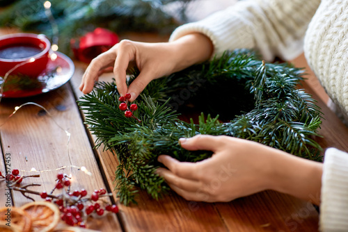 winter holidays, diy and hobby concept - close up of woman with berry decorations making fir christmas wreath at home