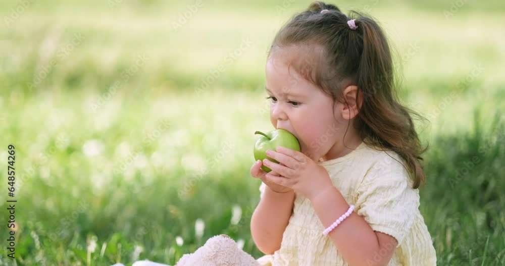 Child little caucasian girl eating apple in park at summer on lunch picnic, kid sitting on grass outside Child little caucasian girl eating apple in park at summer on lunch picnic, kid sitting on grass outside