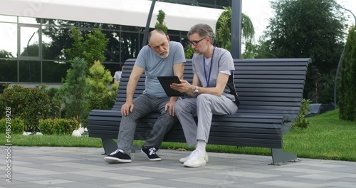 Mature doctor with tablet in hands sits with elderly patient on the bench and consults him. Medic in uniform prescribes medicines to client using digital tablet computer. Medical staff work outdoor.