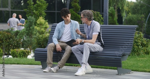 Mature doctor with tablet in hands sits with middle aged patient on bench and consults him. Professional in medical uniform makes diagnosis to client using digital tablet. Medical staff work outdoor.