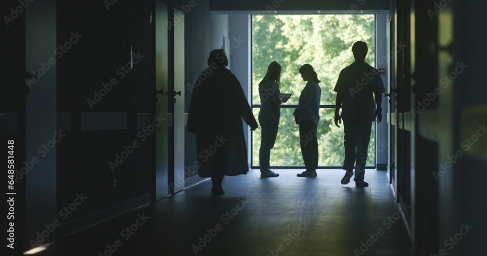 Hospital hallway: Doctors and professional medics walk. Nurse with digital tablet stands with adult woman near window and talks. Medical staff and patients in dark clinic or medical center corridor.
