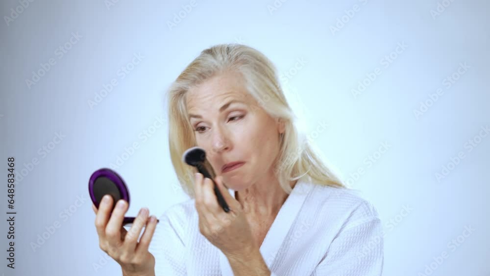 Portrait of mature middle-aged elderly woman wearing robe putting on makeup hygienic look in mirror applying facial powder, face cosmetics lady white studio background Portrait of mature middle-aged elderly woman wearing robe putting on makeup hygienic look in mirror applying facial powder, face cosmetics lady white studio background