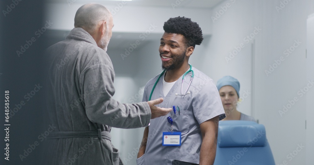 Obraz premium African American doctor consults old patient about his health in clinic corridor. Senior man talks with professional medic. Nurse transports wheelchair down the hospital hallway to medical room.