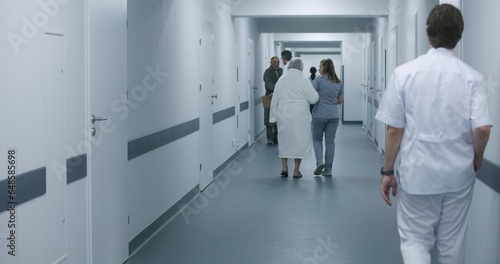 Female doctor, nurse with digital tablet walks along the clinic corridor with elderly woman, helps patient to get to hospital ward after procedures. Medical staff, patients in medical center hallway. © Framestock