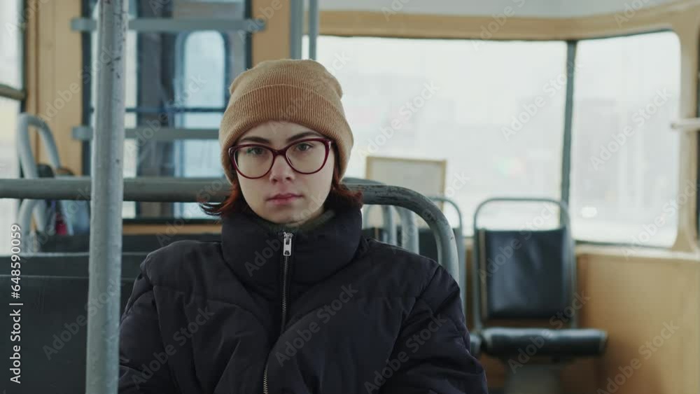Spectacled girl in winter clothes rides in public transport, looks at camera. Portrait of a frozen tired woman in public transport Spectacled girl in winter clothes rides in public transport, looks at camera. Portrait of a frozen tired woman in public transport