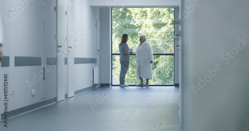 Hospital hallway: Doctors and professional medics walk. Nurse with digital tablet comes to elderly female patient standing near window. Medical staff and patients in clinic or medical center corridor.