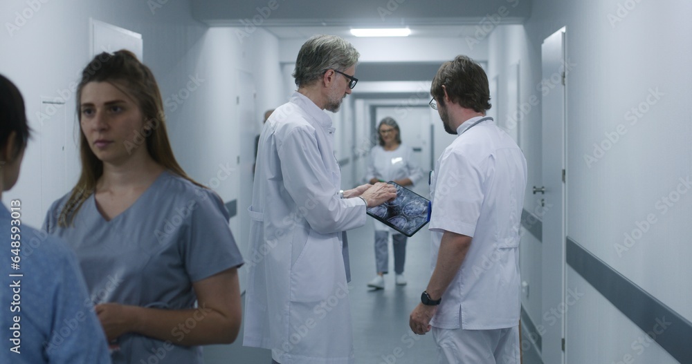 Mature doctor shows MRI, CT brain scan image to colleague using digital tablet. Professional medics stand and walk along the clinic corridor, discuss work. Medical staff, patients in hospital hallway. Mature doctor shows MRI, CT brain scan image to colleague using digital tablet. Professional medics stand and walk along the clinic corridor, discuss work. Medical staff, patients in hospital hallway.