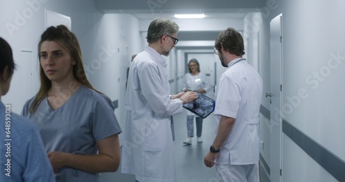 Mature doctor shows MRI, CT brain scan image to colleague using digital tablet. Professional medics stand and walk along the clinic corridor, discuss work. Medical staff, patients in hospital hallway.