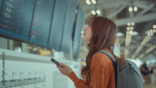 Happy asian woman traveller checking flight schedule departures board in airport terminal hall in front of check in counters. Tourist journey trip concept