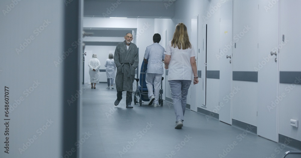 Fototapeta premium Mature doctor stands in clinic corridor with nurse and patient. Professional medic talks with female colleague and elderly woman, uses digital tablet. Medical staff and patients in hospital hallway.