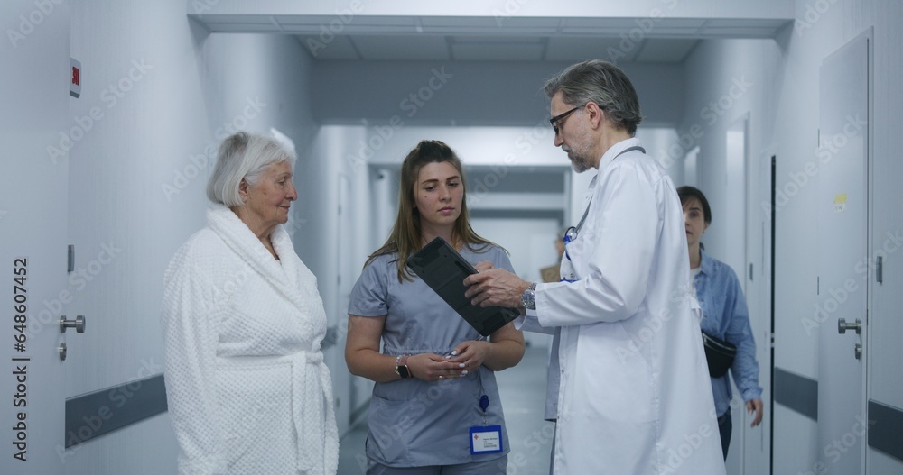 Obraz premium Mature doctor stands in clinic corridor with nurse and patient. Professional medic talks with female colleague and elderly woman, uses digital tablet. Medical staff and patients in hospital hallway.