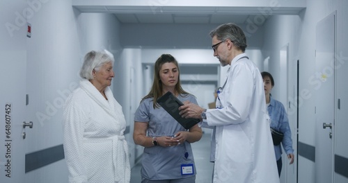 Mature doctor stands in clinic corridor with nurse and patient. Professional medic talks with female colleague and elderly woman  uses digital tablet. Medical staff and patients in hospital hallway.