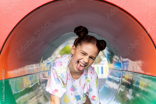фотографія Portrait of a crazy red haired girl with freckles laughing and sticking her tongue out inside a tube in the playground, concept of happiness and joy, carrying a backpack