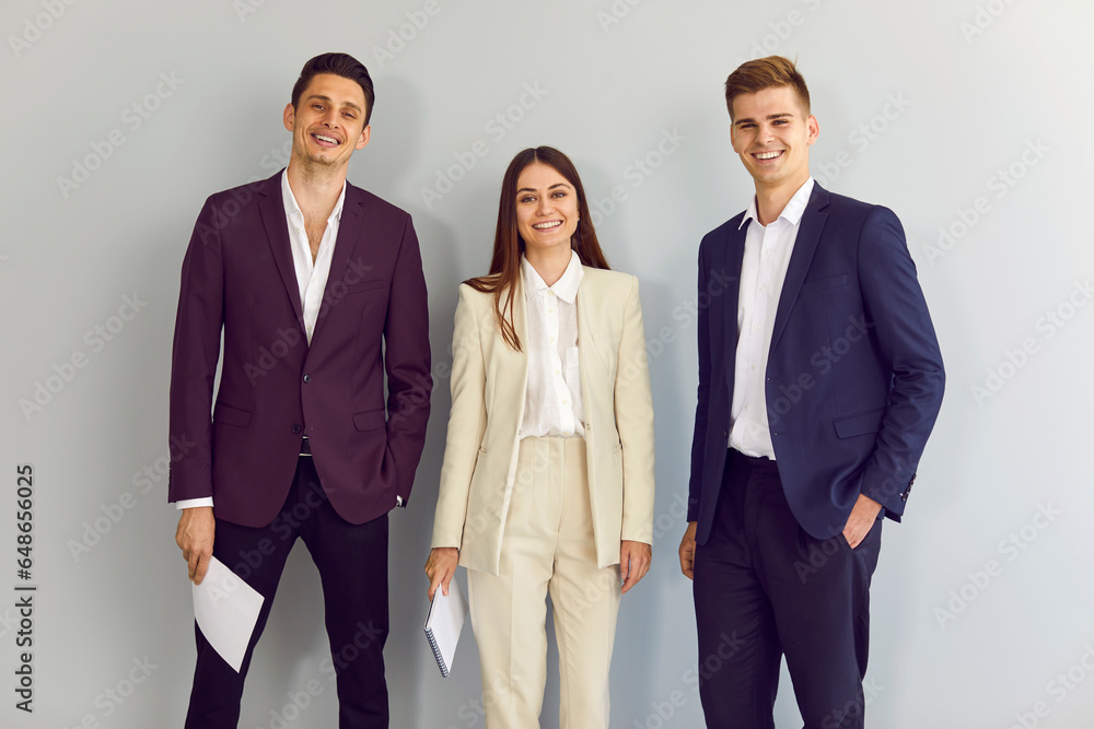 Obraz premium Team of happy young business people standing in the office. Group of two men and one woman wearing elegant, smart, formal dress code blue, purple and beige suits posing by a light grey wall