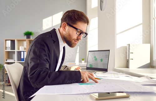 Professional surveyor in suit and glasses sitting at his table in office, working with modern city structure design plans, looking at cadastral maps, and studying plot numbers and boundaries photo