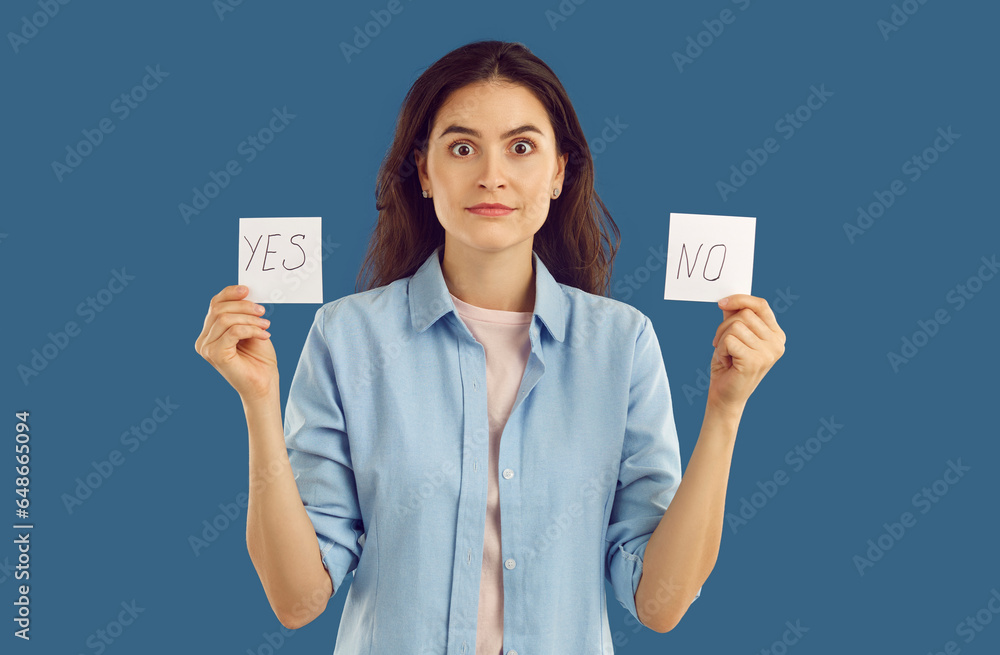 Doubted brunette stunned woman with two cards yes and no looking at camera on blue background. Serious confused undecided woman with face full of doubt looking at camera. Trying to make hard choice. Doubted brunette stunned woman with two cards yes and no looking at camera on blue background. Serious confused undecided woman with face full of doubt looking at camera. Trying to make hard choice.