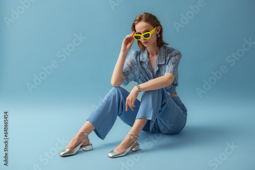 Obraz na plátne Fashionable young woman wearing trendy yellow sunglasses, blue chiffon blouse, trousers, silver ballet flats, posing on blue background