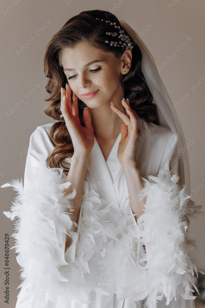 Beautiful young bride with long curly hair in a fashionable white robe in the morning. Morning dressing of the bride. Beautiful young girl in the morning in a white dress. Preparation for the wedding
