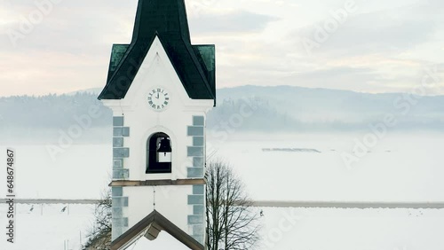 Aerial Orbit Around Top Of Church Standing in Middle Of Fields On Winter Morning. Reveals Bell, Clock, Roof With Fields Covered In Snow With Spruce, Coniferous Forest, Fog, Mist, Road in Background. 