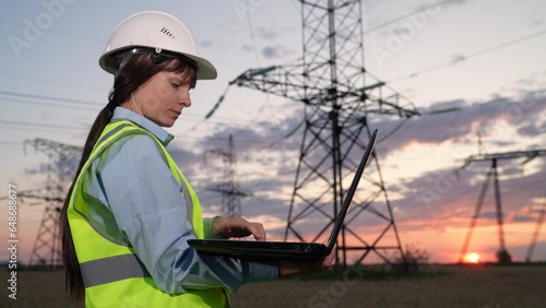 Concentrated employee with helmet works on laptop near power transmission lines