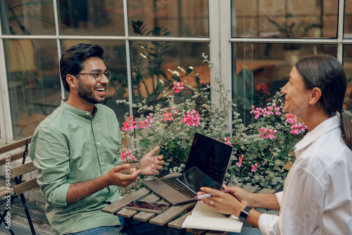 Smiling business colleagues discuss biz issue while use laptop sitting in office terrace