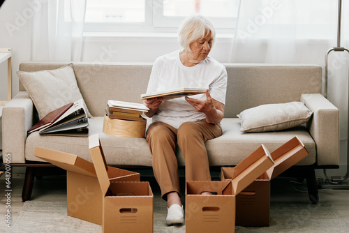 elderly woman sits on a sofa at home with boxes. collecting things with memories albums with photos and photo frames moving to a new place cleaning things and a happy smile. Lifestyle retirement.