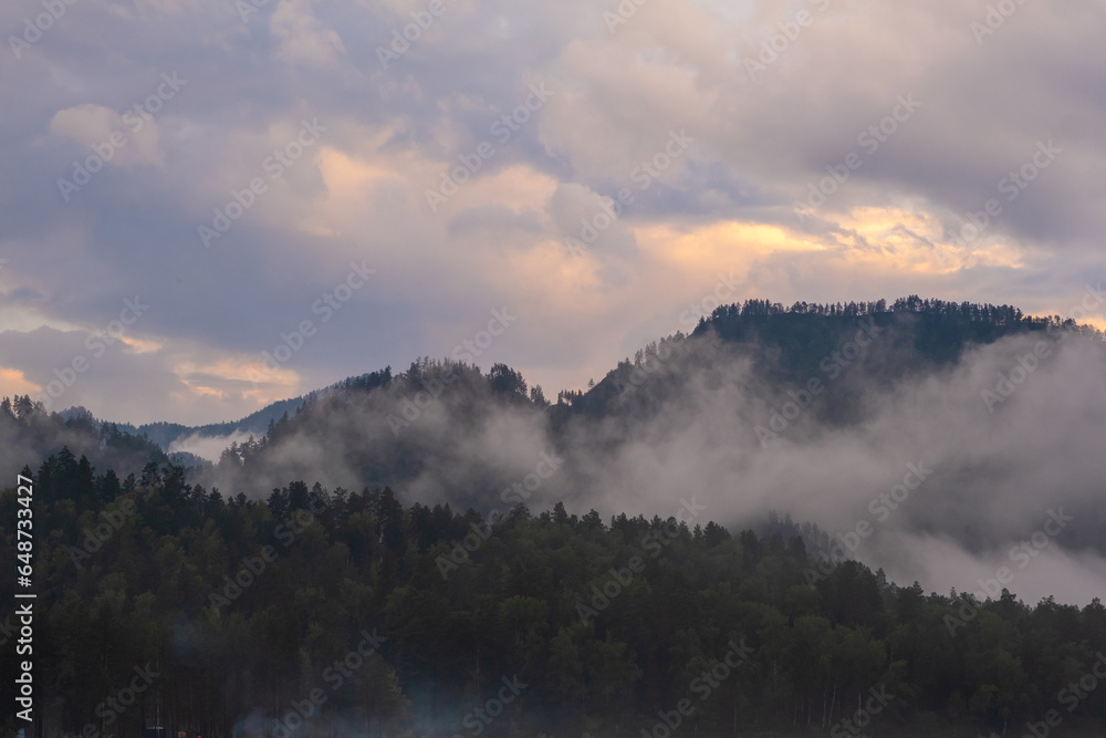Fototapeta premium Mountains and peaks in fog at sunrise or sunset. Sun from behind the mountain. Amazing view of mountains and forest landscape with cloudy skies Altai mountains. Mountains and peaks in fog at sunrise