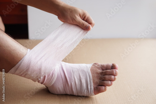 Hand is wrapping bandage around sprain ankle, injury foot treatment. Concept, health problem, accident, first aid. Insurance.         © Sanhanat