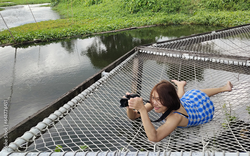 Adorable and smiling Asian woman taking a photo with her camera lying on a net in a river setting surrounded by green vegetation, love of photography, feeling of joy and freedom