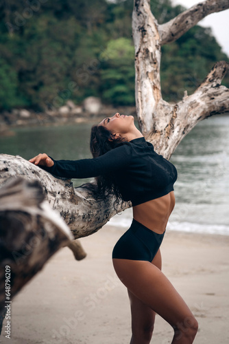 Beautiful girl looking in profile, clouded on a log, in a black section swimsuit with long sleeves