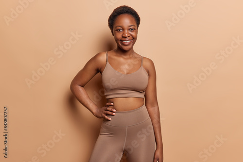 Happy dark skinned African woman keeps hand on waist smiles gladfully has slim body after regular training dressed in activewear smiles pleasantly isolated over brown background being strong