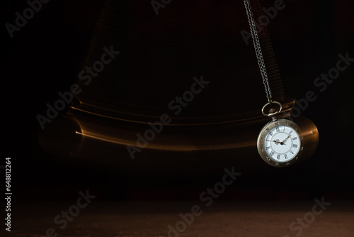 Hypnosis session. Vintage pocket watch with chain swinging over surface on dark background among faded clock faces, magic motion effect photo
