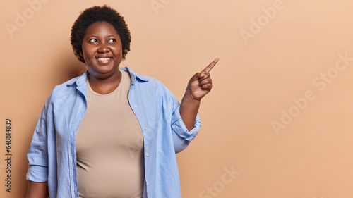 Overweight dark skinned woman with short curly hair points index finger aside shows place for your advertising text wears casual blue shirt isolated over brown background. People and promotion concept