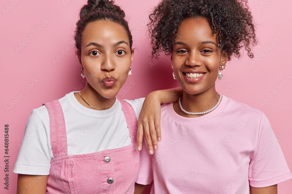 Professional horizontal studio shot of pair of young pretty glad fit African american females wearing casual clothes standing in centre close to each other on pink background without make up Professional horizontal studio shot of pair of young pretty glad fit African american females wearing casual clothes standing in centre close to each other on pink background without make up