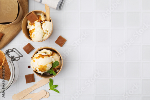 Scoops of ice cream with caramel sauce, mint leaves and candies on white tiled table, flat lay. Space for text