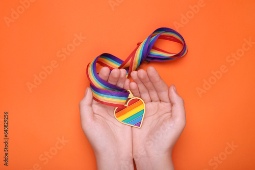 Woman holding rainbow ribbon with heart shaped pendant on orange background, top view. LGBT pride