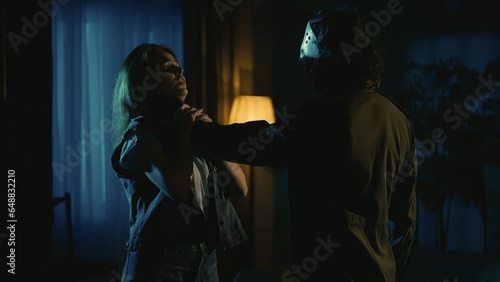 Man in mask and girl standing in the dark apartment living room, maniac holding girl by the neck and strangling her.