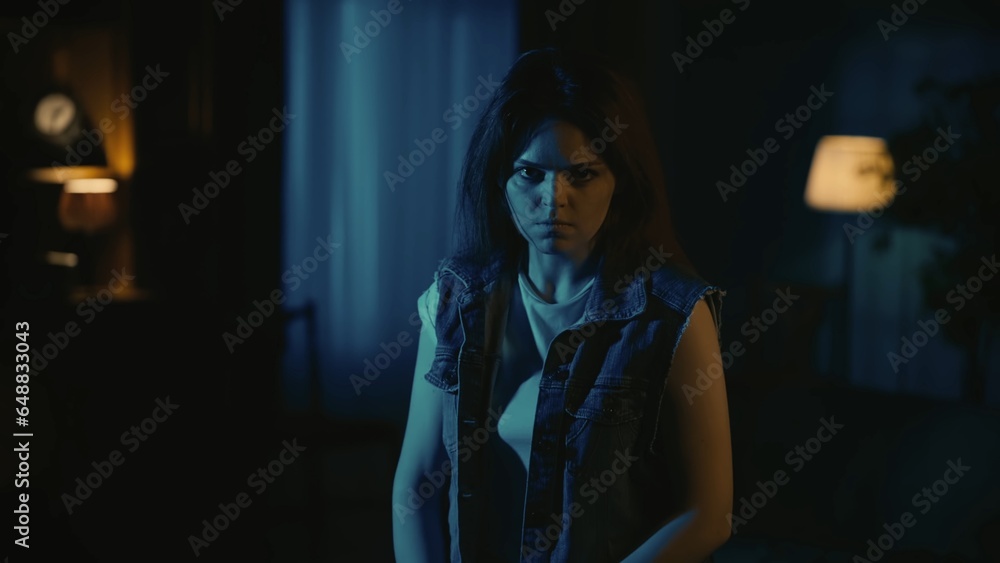 Girl in casual clothing with angry face expression looking at the camera standing in the dark living room, tense atmosphere. Girl in casual clothing with angry face expression looking at the camera standing in the dark living room, tense atmosphere.