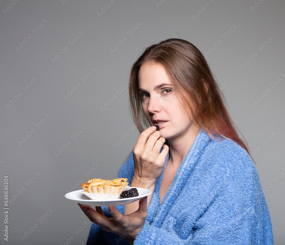 sallow unhealthy looking woman eating fruit and pastry wearing a blue bathrobe looking at the camera. Diet and self care concept image. sallow unhealthy looking woman eating fruit and pastry wearing a blue bathrobe looking at the camera. Diet and self care concept image.