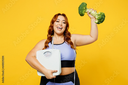 Young happy chubby overweight plus size big fat fit woman wear blue top warm up training hold measure tape, broccoli, scales isolated on plain yellow background studio home gym. Workout sport concept. photo