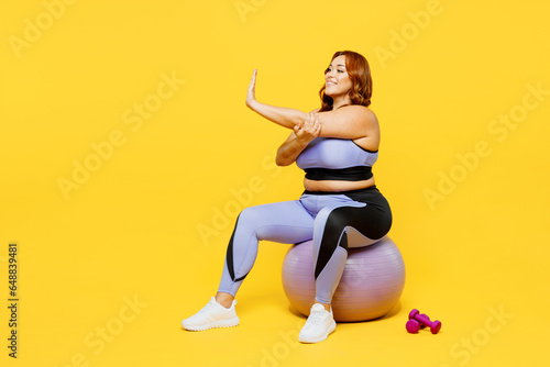 Full body young chubby plus size big fat fit woman wear blue top warm up train sit on fit ball do stretch exercise for hand isolated on plain yellow background studio home gym. Workout sport concept.