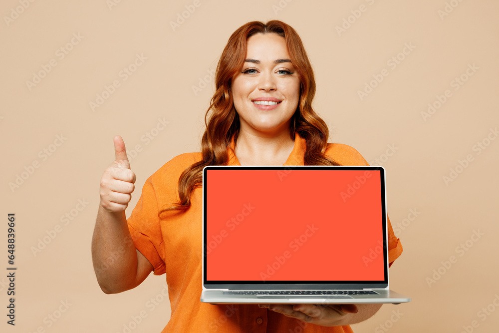 Young ginger chubby overweight IT woman wears orange shirt casual clothes hold use work on blank screen laptop pc computer show thumb up isolated on plain pastel beige background. Lifestyle concept. Young ginger chubby overweight IT woman wears orange shirt casual clothes hold use work on blank screen laptop pc computer show thumb up isolated on plain pastel beige background. Lifestyle concept.