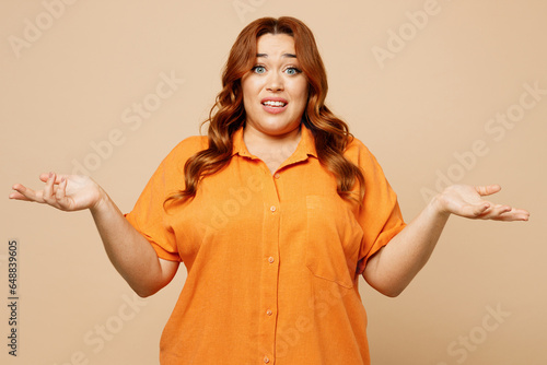 Young sad ginger chubby overweight woman wear orange shirt casual clothes spread hands shrugging shoulders look puzzled, have no idea isolated on plain pastel light beige background studio portrait photo