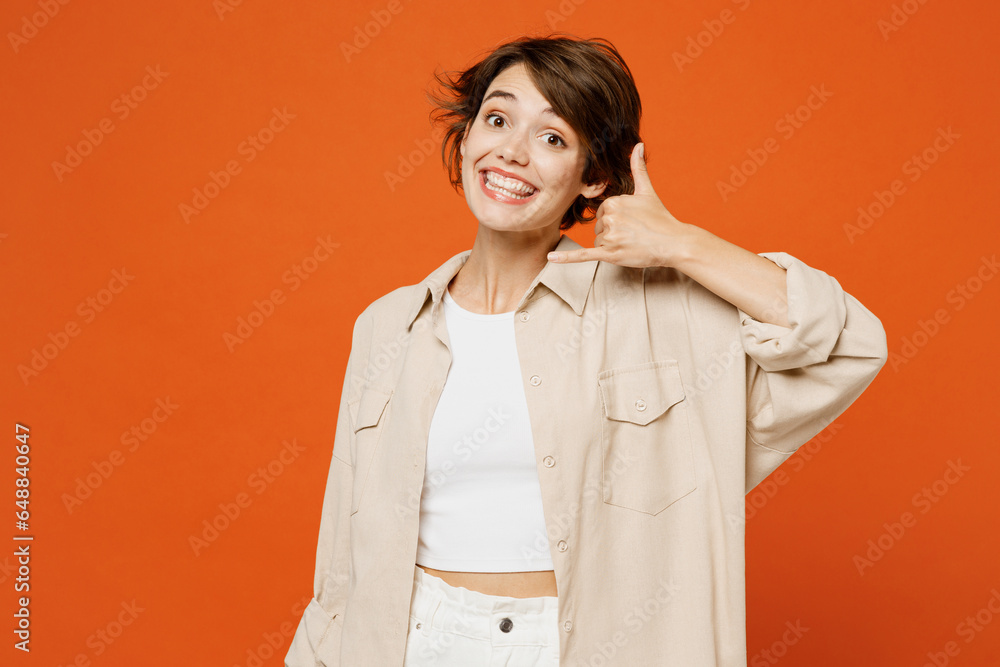 Young smiling cheerful happy caucasian woman she wear beige shirt casual clothes doing phone gesture like says call me back isolated on plain orange red background studio portrait. Lifestyle concept. Young smiling cheerful happy caucasian woman she wear beige shirt casual clothes doing phone gesture like says call me back isolated on plain orange red background studio portrait. Lifestyle concept.